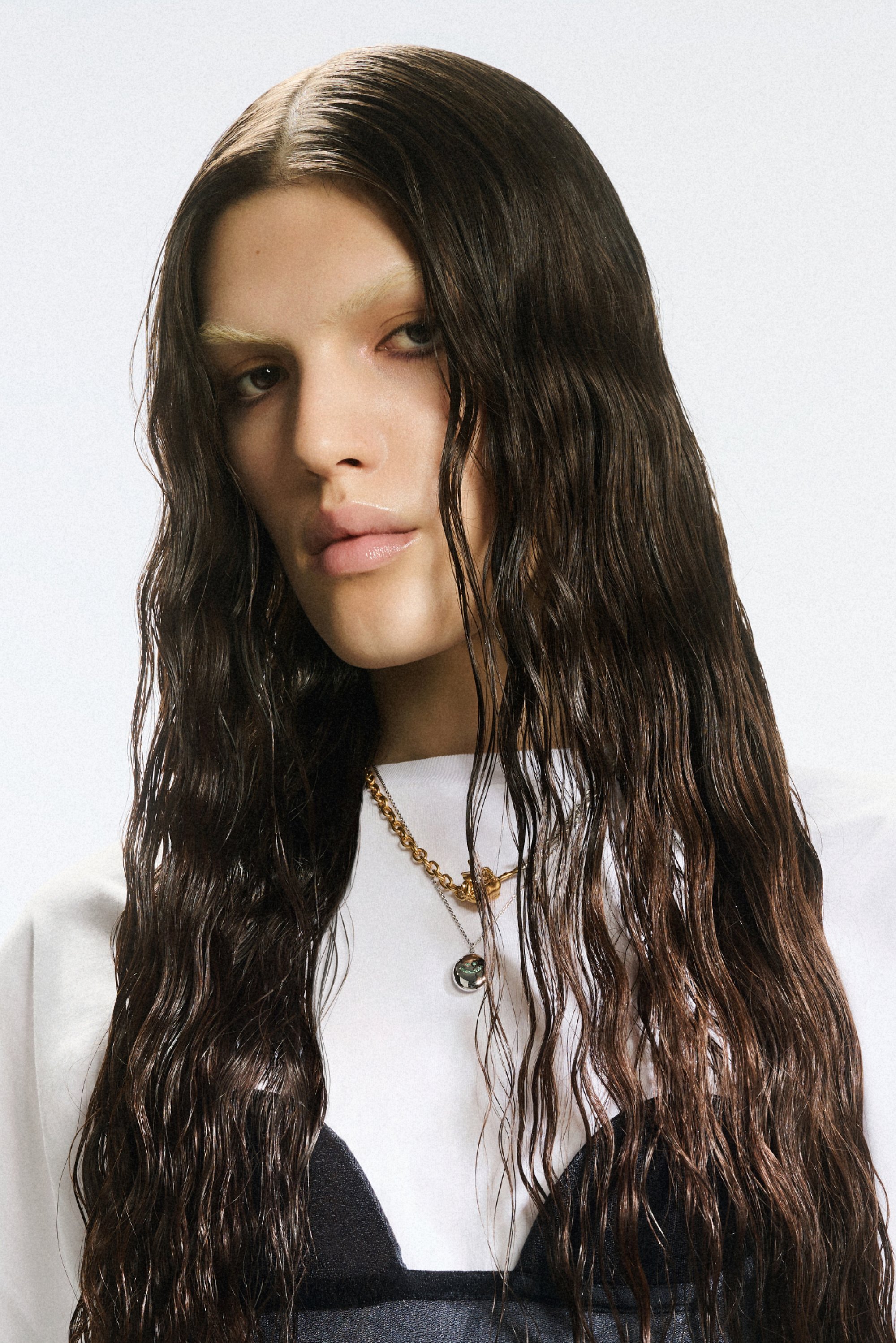 Model met lang, nat haar, in een wit shirt en gelaagd zwart topje, versierd met een gouden ketting en hanger, kijkt rechtstreeks in de camera.