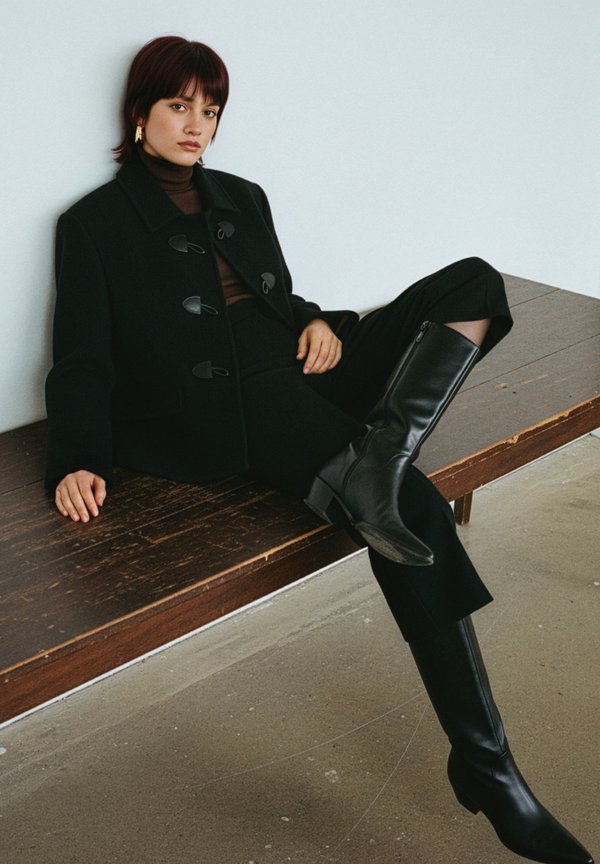 Svart ullkappa med toggelstängningar, brun polotröja, skräddarsydda svarta byxor och knähöga svarta läderstövlar. Träbänk och neutral bakgrund.
