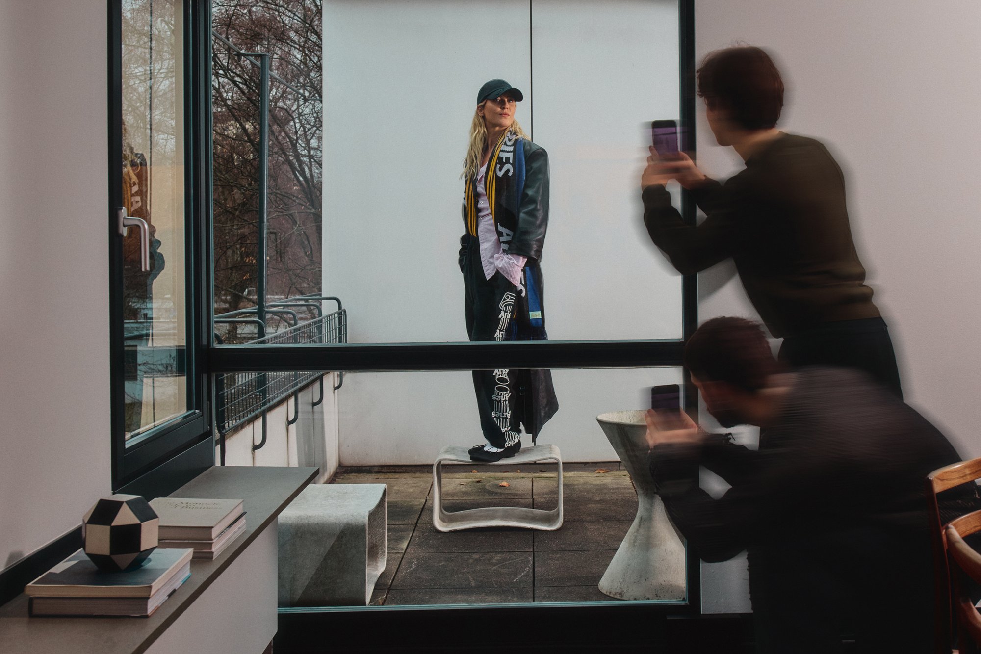 Una modella è in piedi su uno sgabello all'aperto, indossando un abbigliamento alla moda. Una persona cattura il momento con un telefono attraverso la porta di vetro.