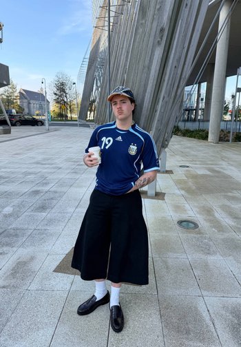Joven con camiseta deportiva azul y gorra se apoya en vigas de madera al aire libre, sosteniendo una taza de café, lleva pantalones negros de pierna ancha y mocasines.