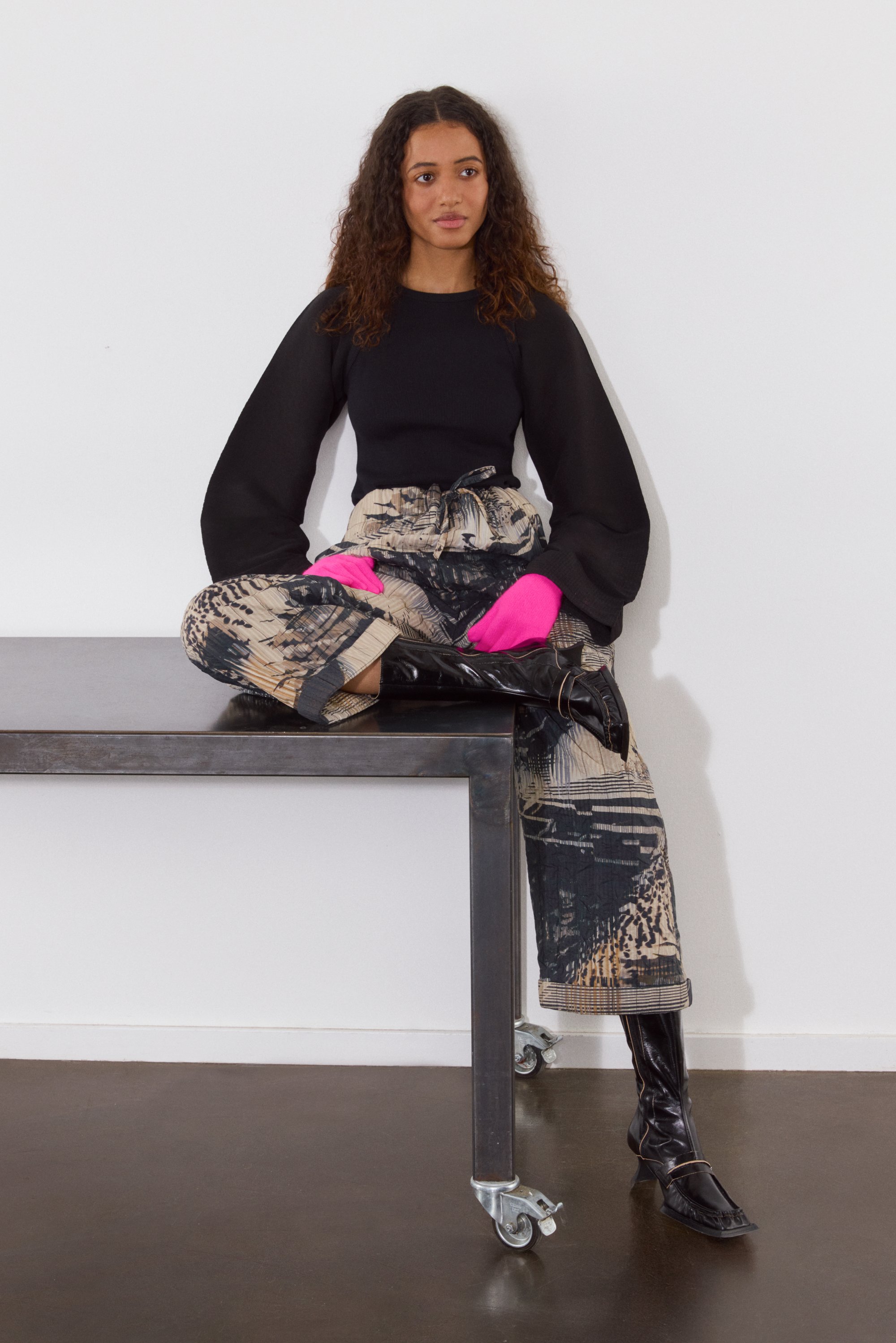 Una joven con cabello rizado se sienta sobre una mesa, vistiendo una blusa negra, pantalones con estampado y guantes rosas brillantes, con un fondo minimalista.