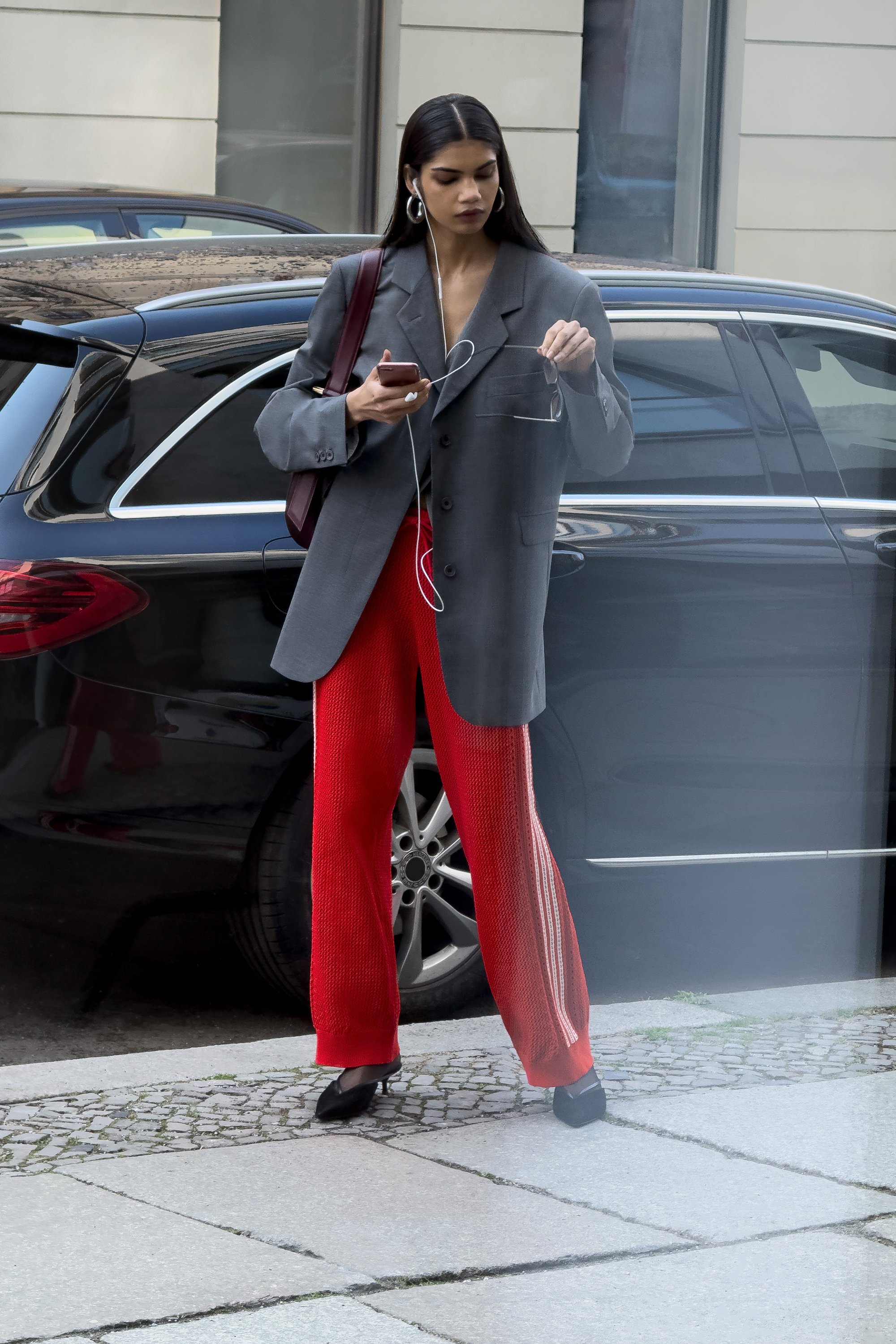 Een vrouw in een oversized grijze blazer en rode broek staat bij een zwarte auto, houdt een telefoon vast en past haar zonnebril aan, met oordopjes in.