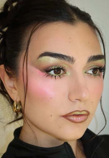 Frau mit metallisch goldem Lidschatten und Lippenstift, rosa Rouge, dichten Augenbrauen, langen Wimpern, goldenen Creolen und dunkel zurückgebundenen Haaren.