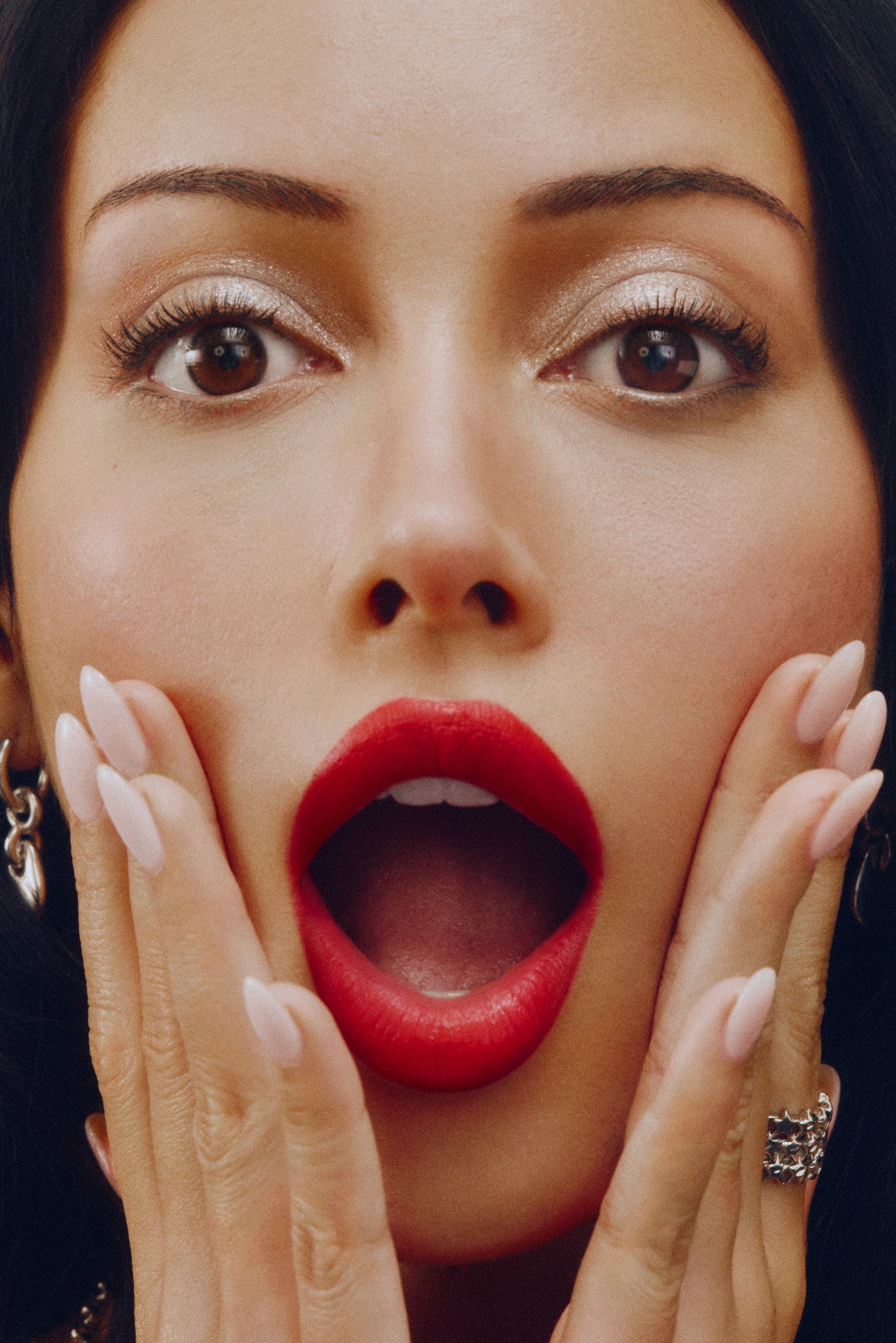 Primer plano de un rostro con cabello oscuro y expresión de sorpresa. Presenta labios rojos intensos, uñas manicura de color rosa claro y sombra de ojos brillante.