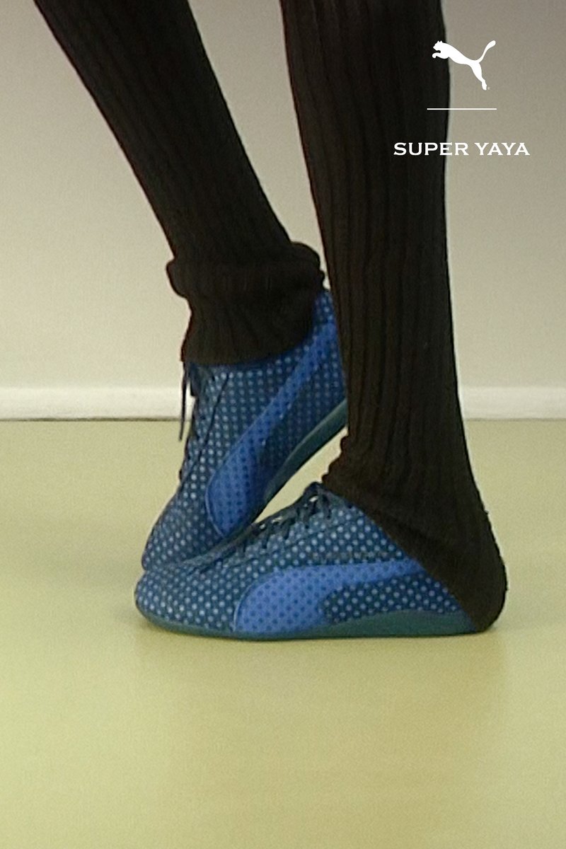 Person trägt schwarze gerippte Hose und blaue Puma Super Yaya Sneaker, steht auf hellem Boden.