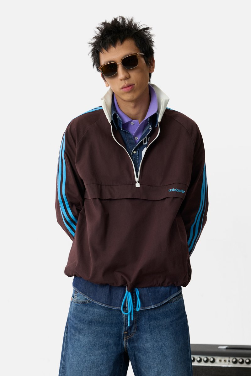 Chaqueta marrón de pullover con rayas azules en las mangas, cuello con cremallera y dobladillo con cordón. Lleva puesta sobre una camisa de mezclilla azul y un polo morado.