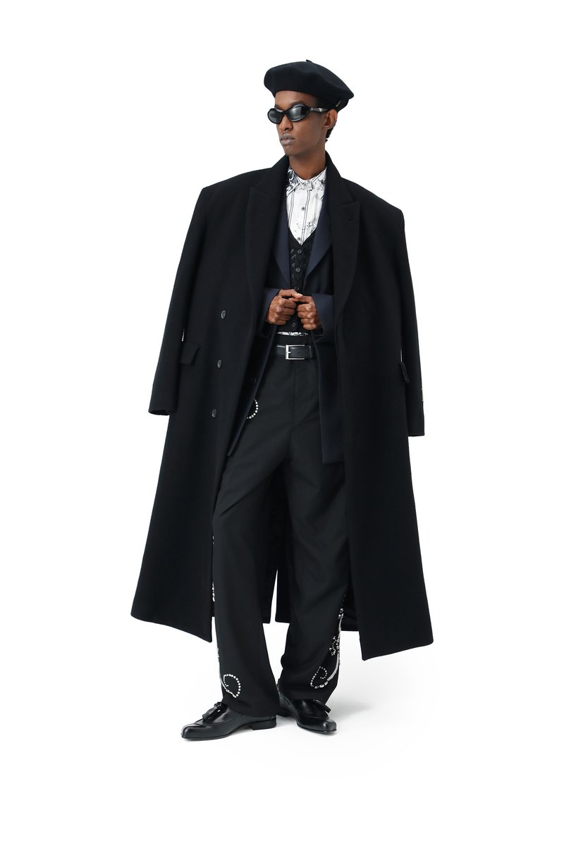 Una modella si erge con sicurezza indossando un lungo cappotto nero, pantaloni alla moda, una camicia a fantasia, occhiali da sole e un basco, emanando un aspetto elegante e moderno.