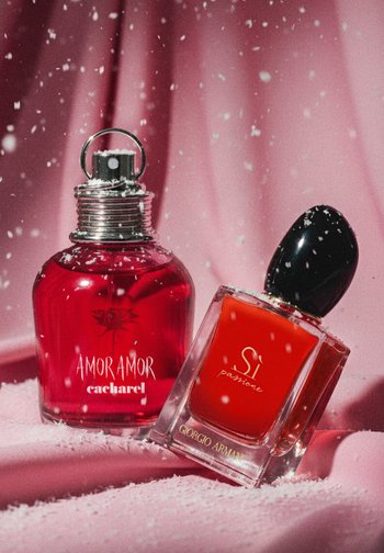 Dwa flakony perfum na różowym tle: okrągły, ciemnoczerwony flakon z metalowym spryskiwaczem oznaczony "Amor Amor" oraz prostokątny przezroczysty flakon z czarną nakrętką oznaczony "Si."