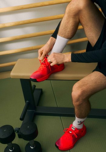 Person sitting on a bench tying red athletic shoes, wearing white socks and black athletic shorts, with dumbbells on the floor nearby.