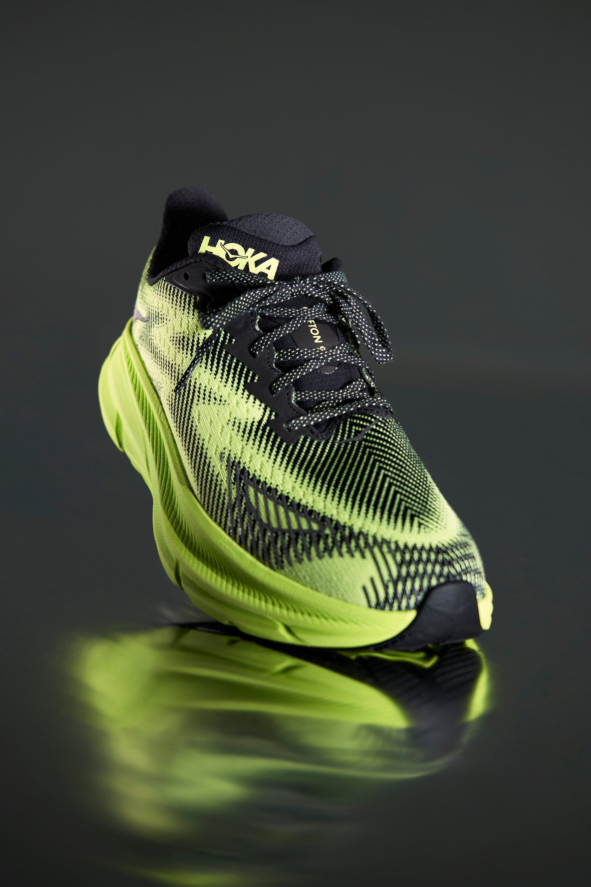Zapatilla de correr HOKA en verde y negro con un diseño texturizado, mostrada en un ángulo sobre una superficie reflectante, resaltando su forma y colores.
