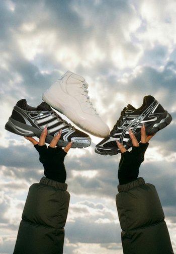 Three pairs of trainers held up: a white high-top, a black and silver low-top, and a black and grey sneaker. Cloudy background.