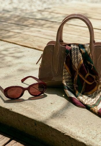 Hnědé sluneční brýle, béžová kabelka a vzorovaný šátek leží na osvětleném betonovém povrchu s dřevěnými prkny v pozadí.