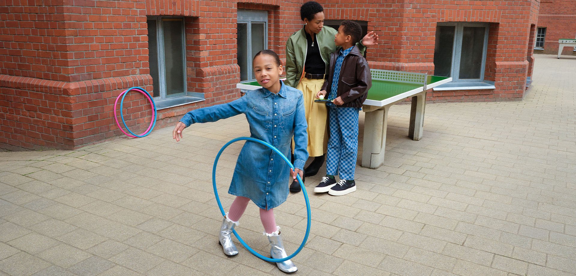 Niña con vestido vaquero jugando con un hula hoop azul al aire libre, otros dos están junto a una mesa de ping-pong junto a un edificio de ladrillo.