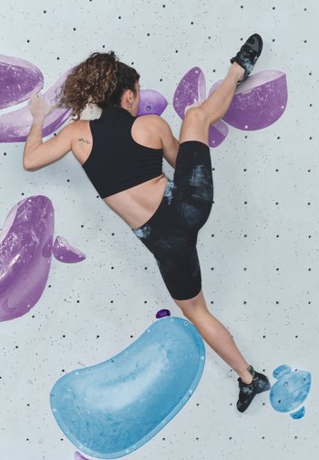 Escenario de escalada en roca con una persona en una pared blanca texturizada con presas moradas y azules. El individuo lleva un top corto negro y shorts.