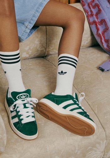 Grüne Wildledersneaker mit weißen Streifen, kombiniert mit gestreiften weißen Socken, ruhen auf einem strukturierten beigen Sofa. Heller und lässiger Schuhstil.