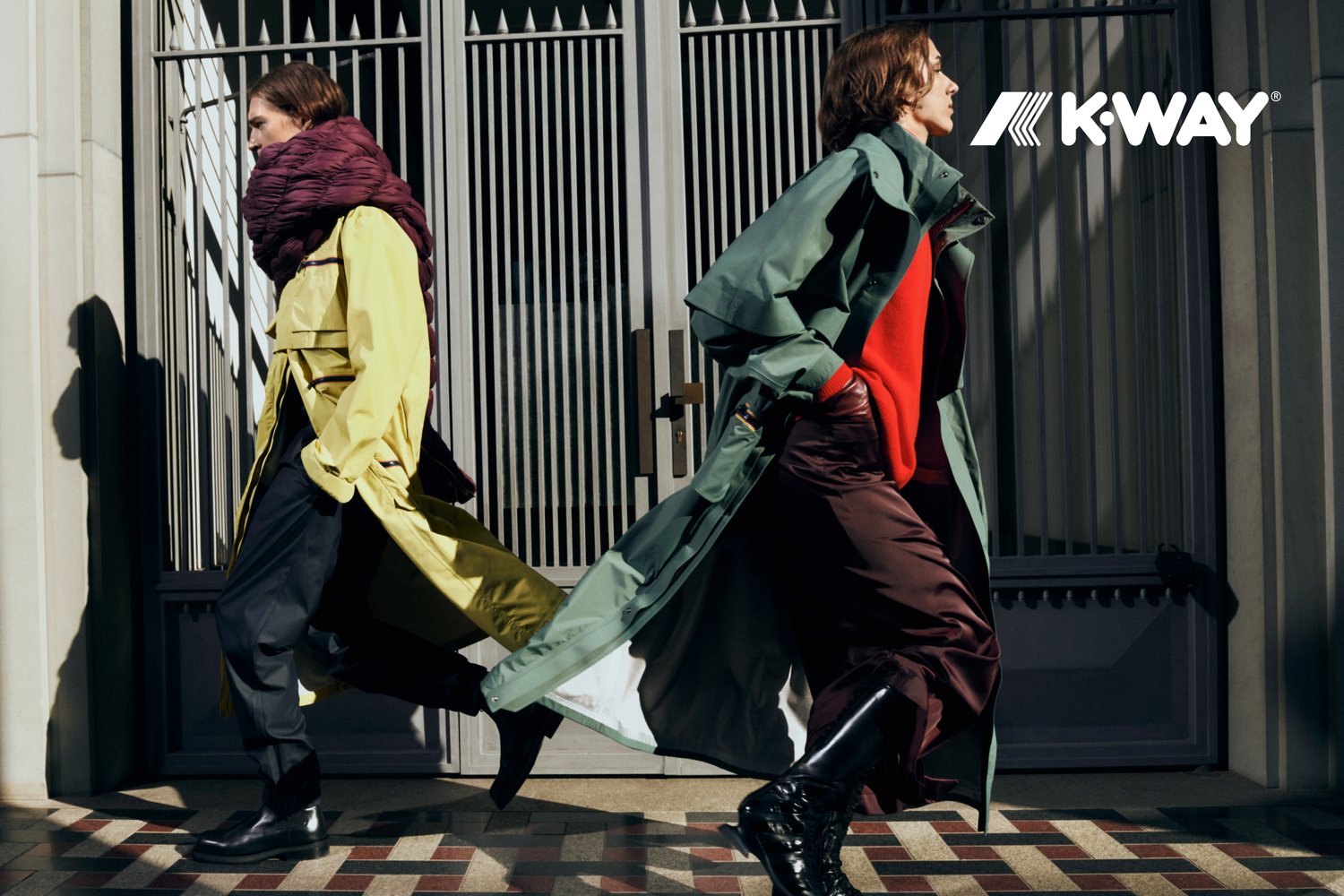 Zwei Models laufen in einer Stadtkulisse, bekleidet mit einem gelben Trenchcoat und einem grünen Mantel, ergänzt durch einen auffälligen roten Pullover und dunkle Hosen.
