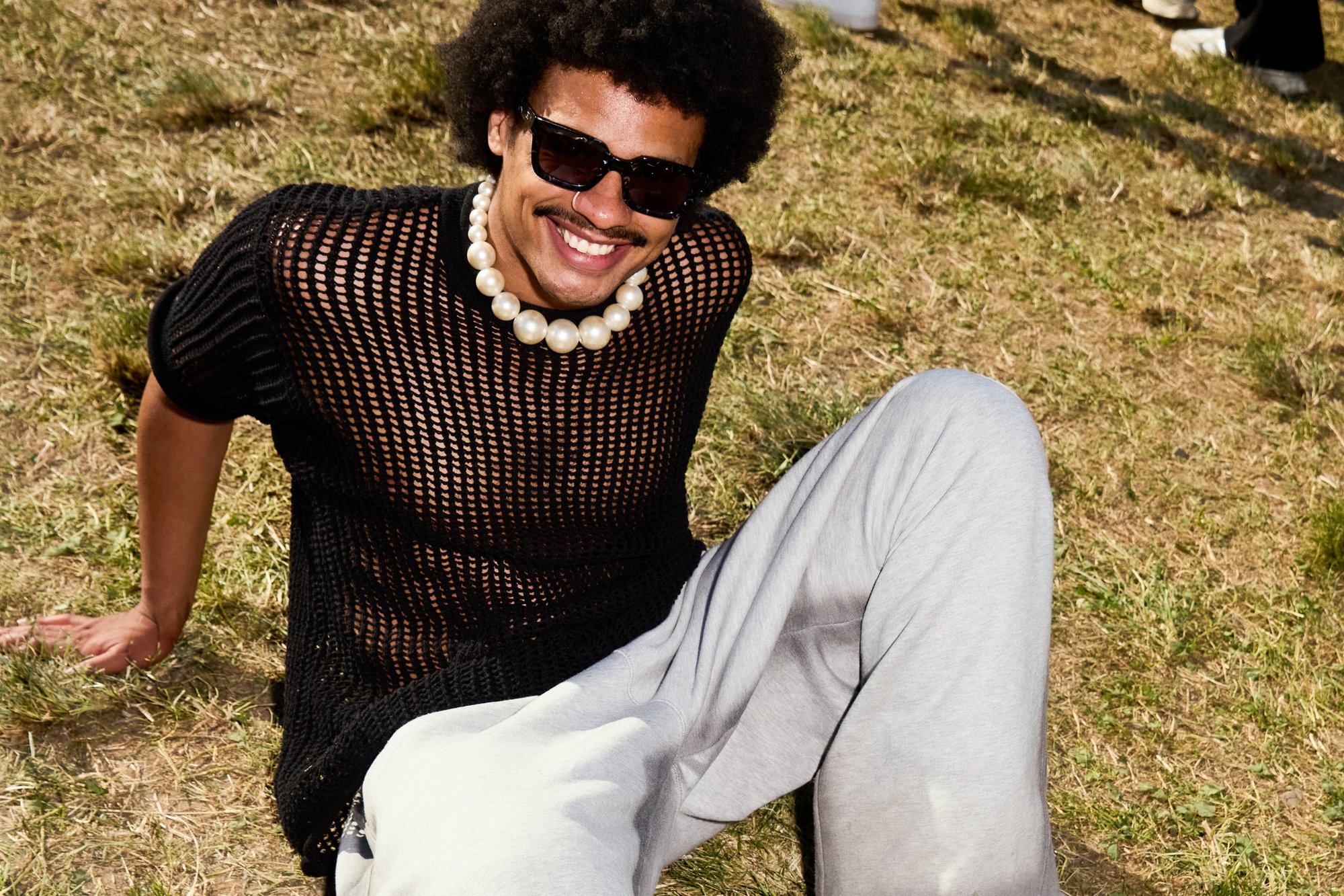 Un pull en maille noir, un pantalon de survêtement gris clair et un grand collier en perles. Assise sur l'herbe avec des lunettes de soleil, affichant une posture détendue et un sourire.