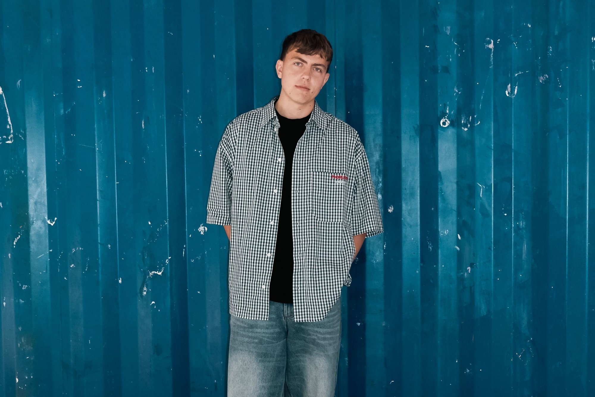 Ung man med kort brunt hår som bär en rutiga skjorta och jeans, stående med händerna bakom ryggen mot en texturerad blå vägg.