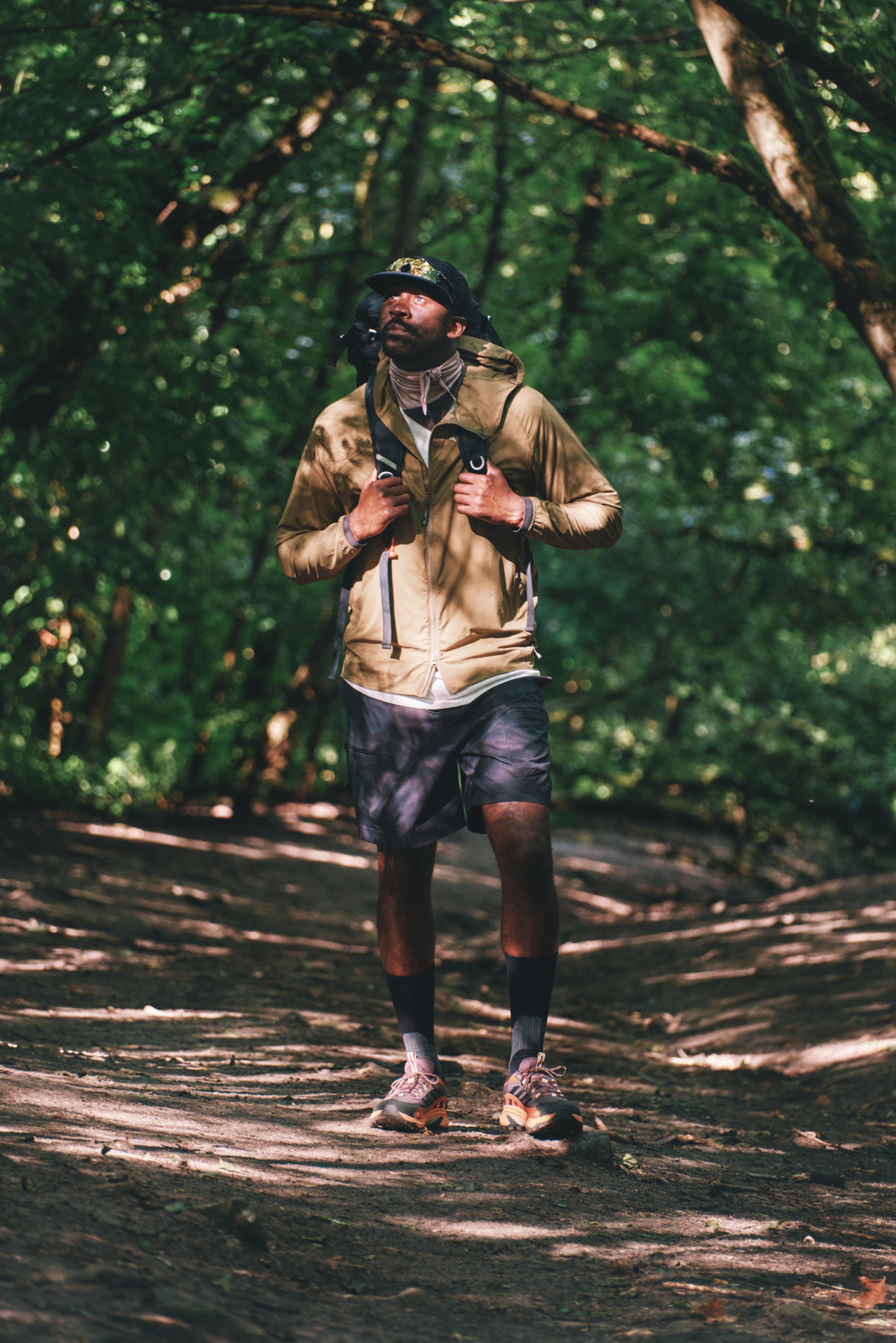 Lichtgewicht tan jacket, grijze shorts en wandelschoenen gedragen door een persoon die op een dirt pad loopt, omringd door groene bladeren.
