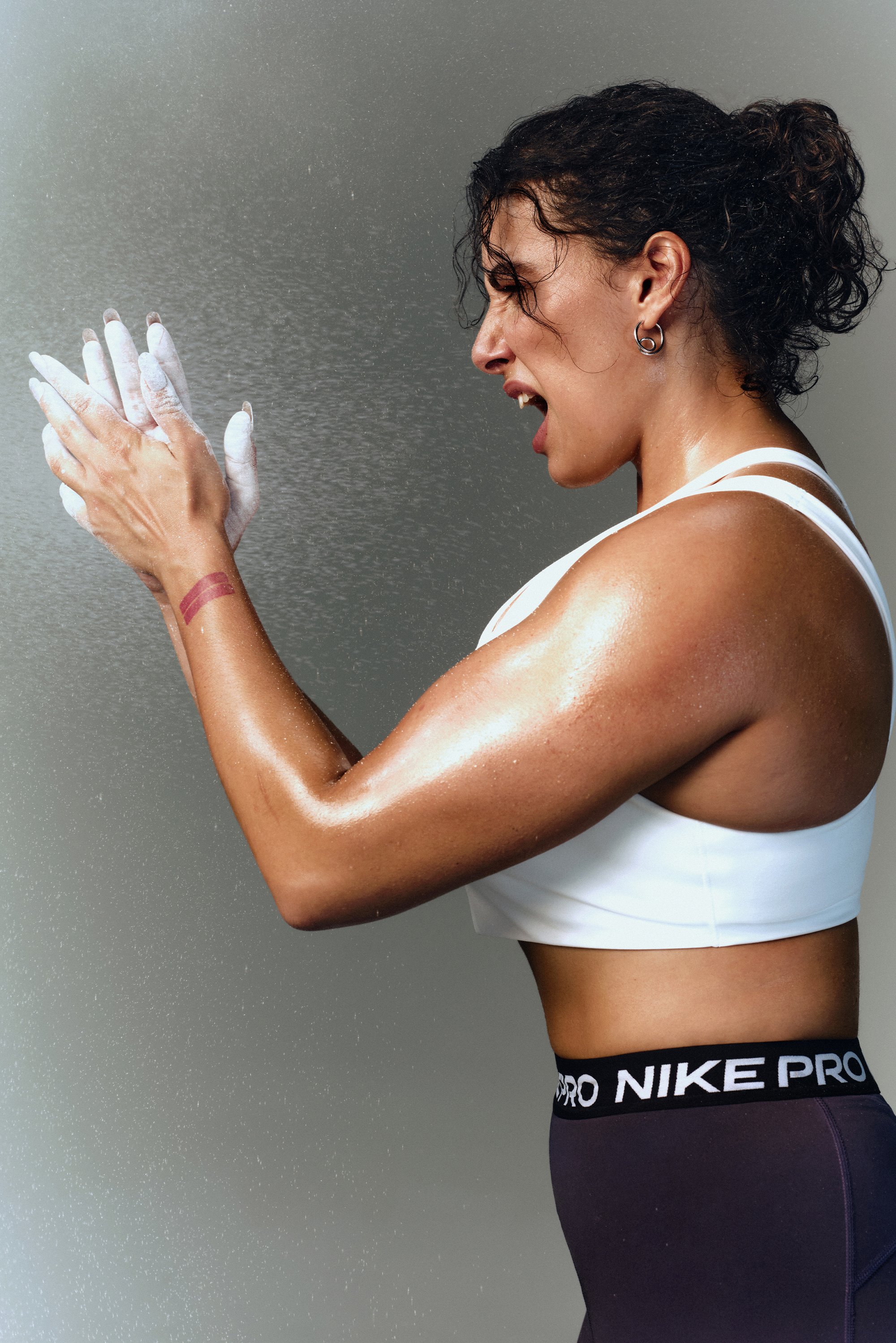 A muscular woman prepares for a workout by applying chalk to her hands, displaying focused intensity and determination in her expression.