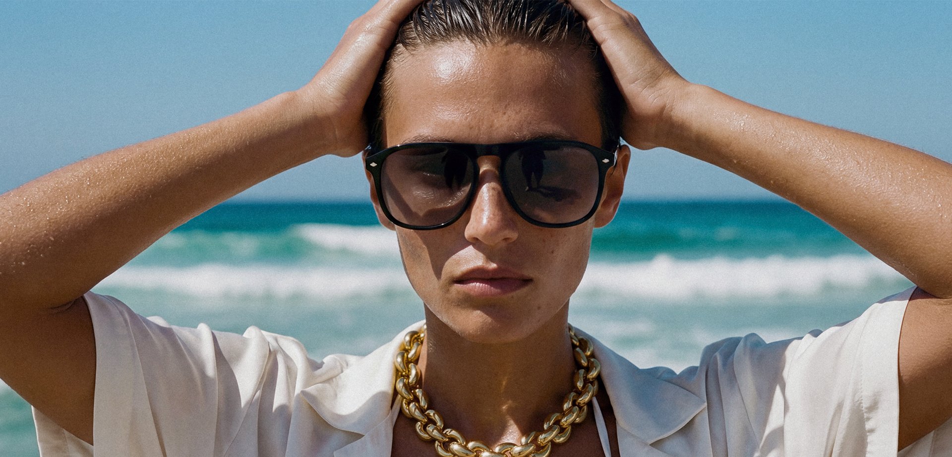 Άτομο που φοράει μεγάλα μαύρα γυαλιά ηλίου και χρυσή αλυσίδα κρατάει το κεφάλι με τα δύο χέρια με φόντο τα κύματα του ωκεανού.