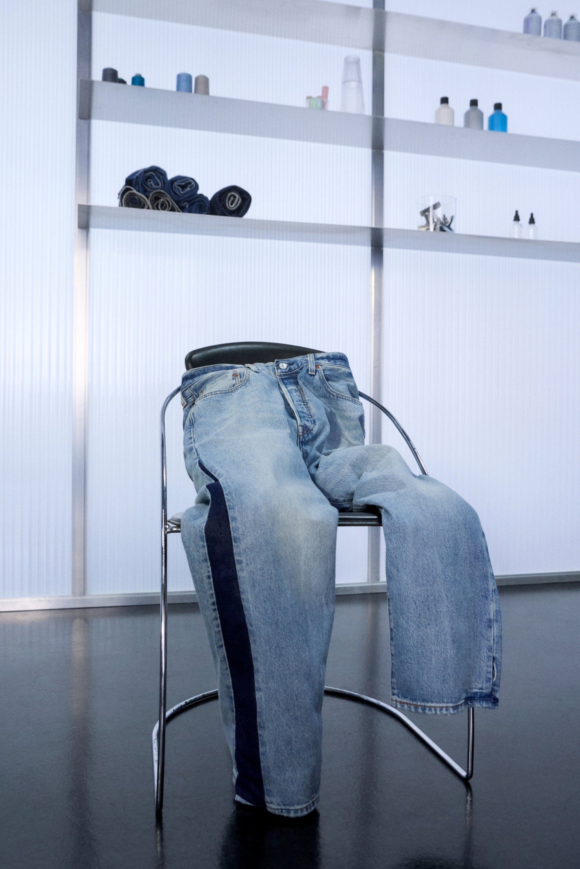 Denim jeans gepresenteerd op een moderne chroomstoel, met een minimalistische achtergrond waarin planken met opgerolde handdoeken en flessen zijn te zien.