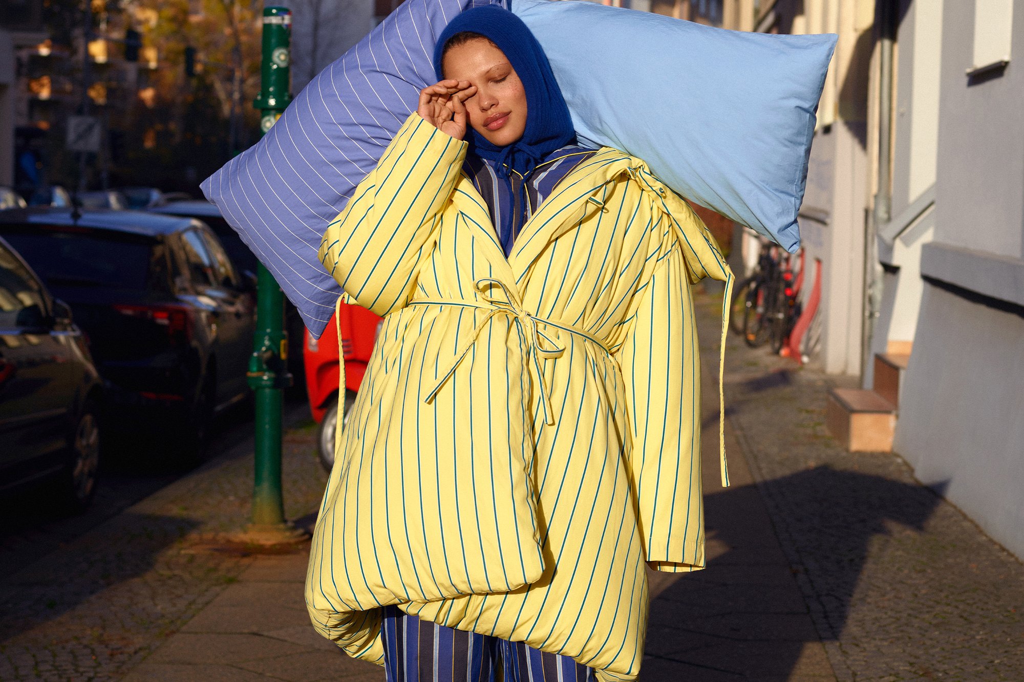 Una persona in un cappotto a righe gialle e blu porta due cuscini, uno blu e uno a righe, su un marciapiede urbano illuminato dal sole.