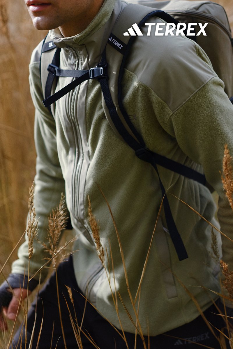 Fleece jacket in light green with a zip front, high collar, and reinforced shoulder sections. Adjustable straps and a rucksack are visible.