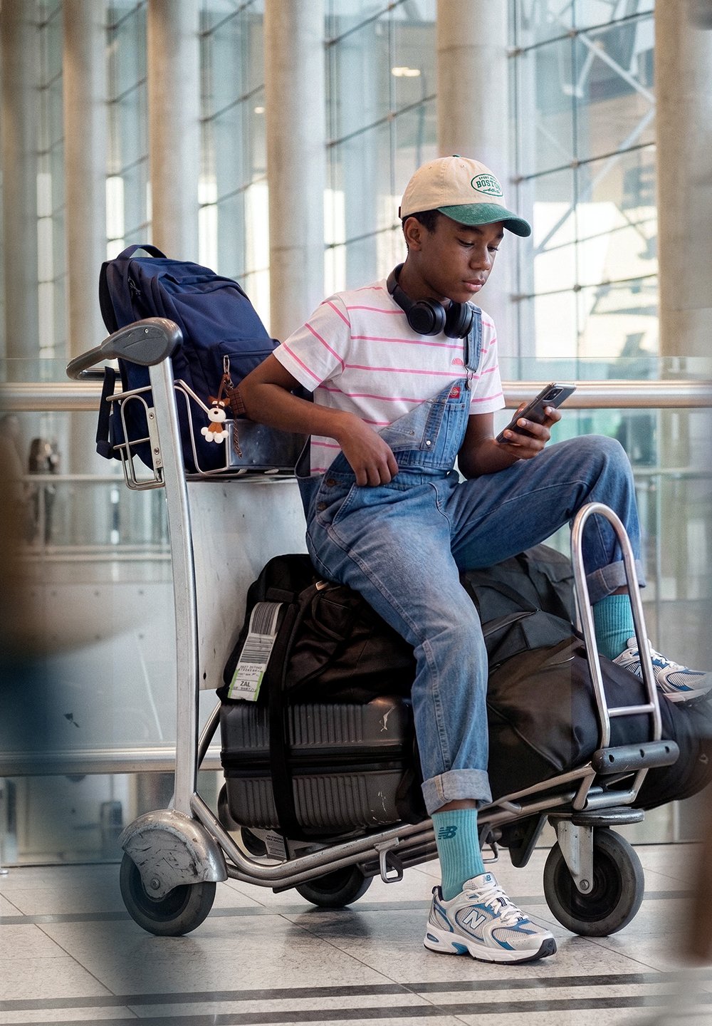 Teenager mit Kappe und Kopfhörern sitzt auf einem Gepäckwagen am Flughafen und schaut auf sein Handy, umgeben von Koffern und Rucksäcken.