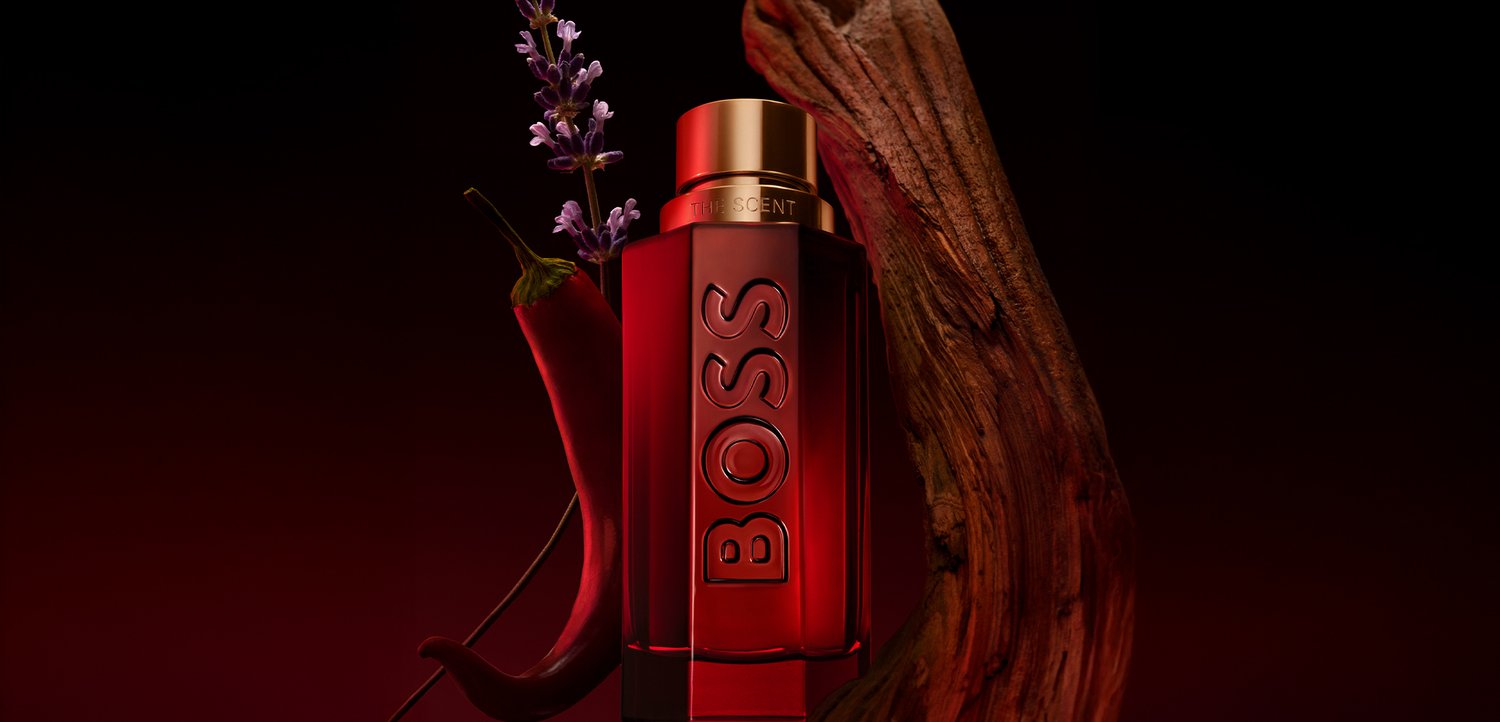 Bottiglia di profumo BOSS rossa con tappo dorato, posizionata accanto a un peperoncino rosso, un fiore viola e legno strutturato su uno sfondo scuro.