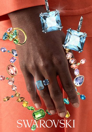 Hand mit großem blauem Edelsteinring, umgeben von buntem Edelsteinschmuck einschließlich Ohrringen, Halskette und Ring auf orangefarbenem Stoffhintergrund.
