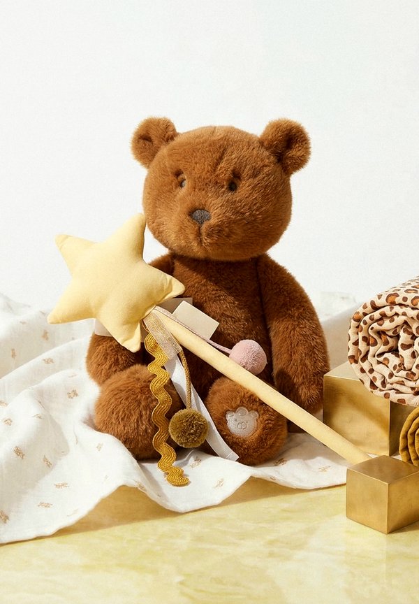 Brown teddy bear holding a yellow star wand sitting on a white cloth, next to rolled patterned fabrics and gold cube blocks.