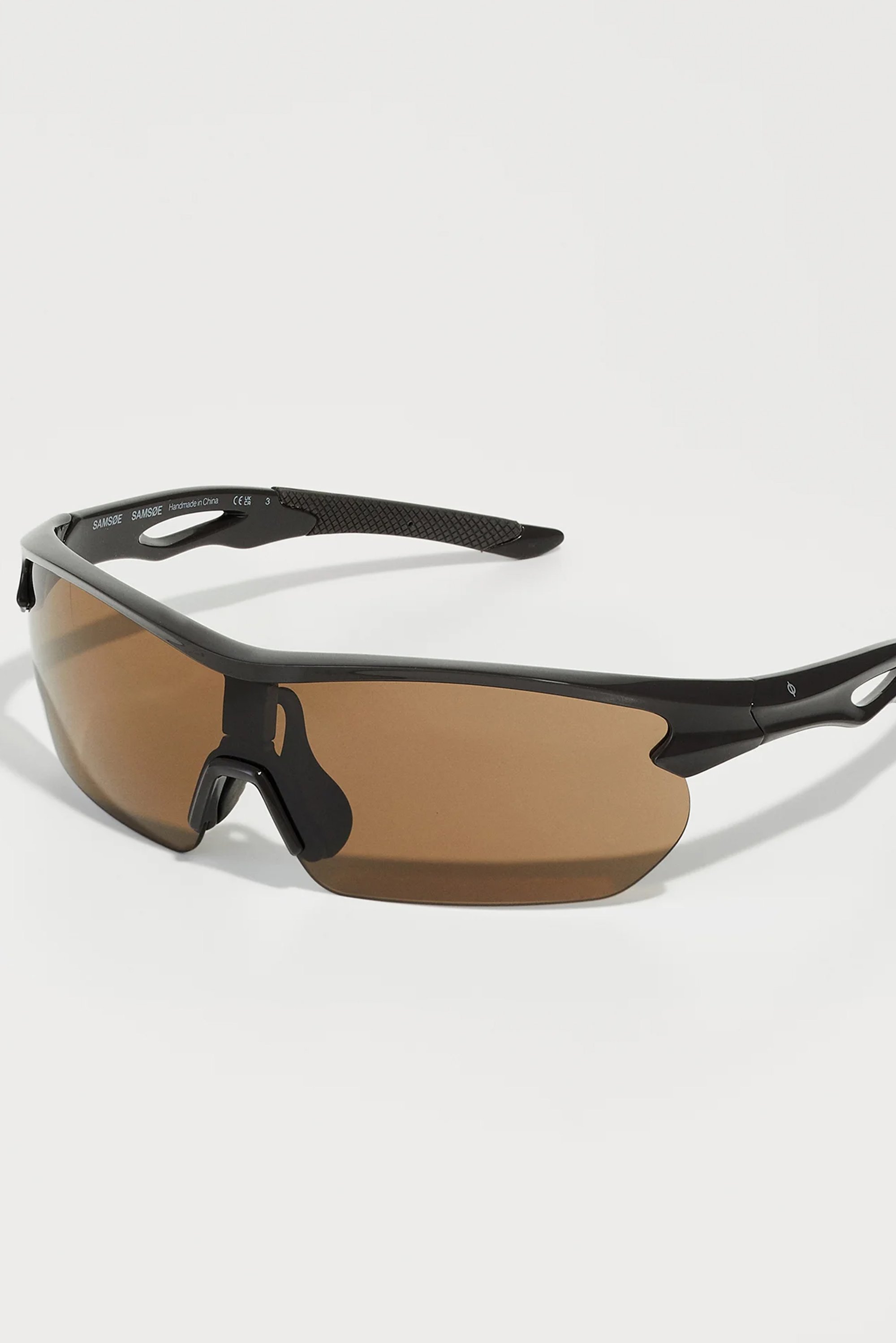 Gafas de sol deportivas tintadas de marrón con un elegante marco negro, lentes angulares, almohadillas nasales de goma y empuñaduras laterales de goma texturizada para un uso seguro.