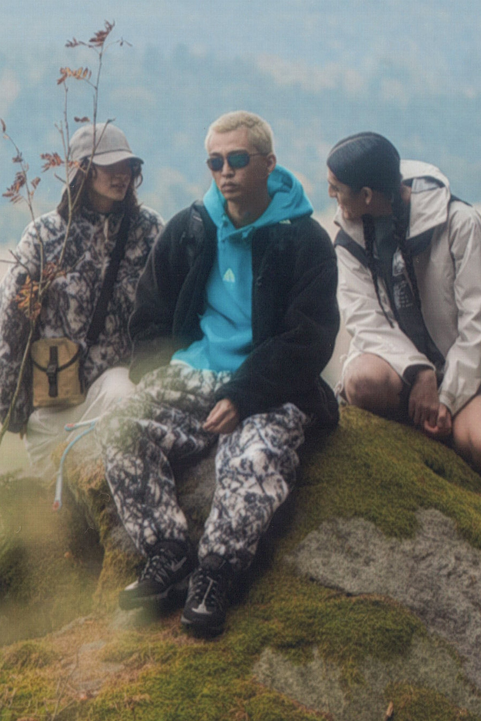 Tres amigos están sentados en una roca cubierta de musgo en un bosque, vistiendo ropa de exterior colorida y compartiendo una conversación ligera.