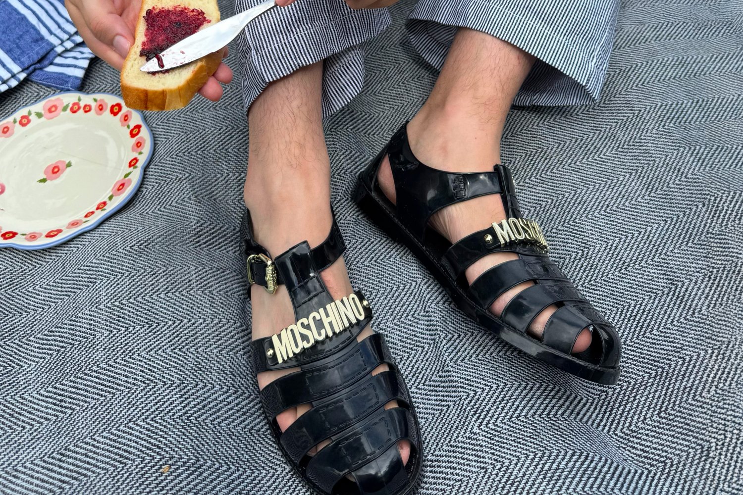 Sandali neri in plastica con design a fori, caratterizzati dal branding dorato "MOSCHINO". Nel background si vedono pantaloni a righe e un piatto con del cibo.