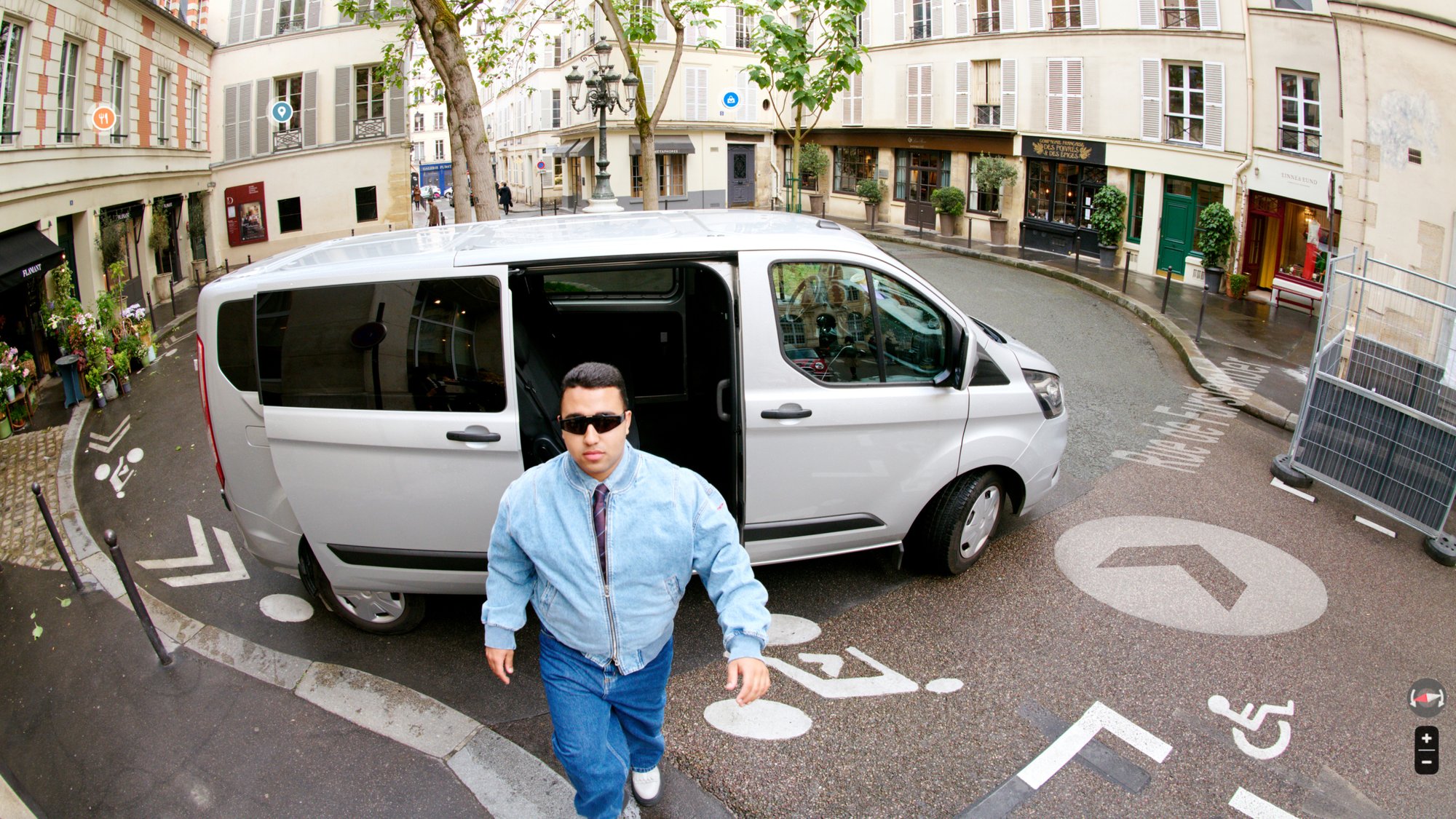 Un hombre con gafas de sol sale de una furgoneta plateada en una calle estrecha con tiendas y árboles, junto a símbolos de accesibilidad en la acera.
