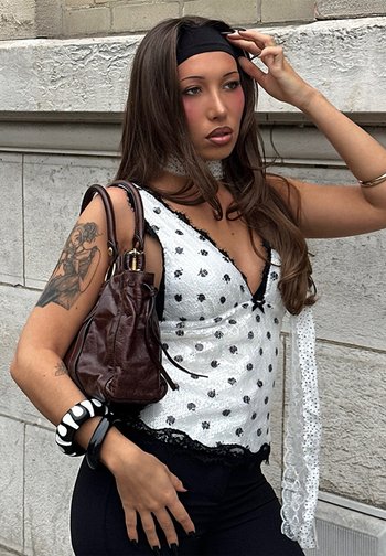 Jeune femme aux longs cheveux bruns porte un haut blanc à pois, un bandeau noir, un pantalon noir et porte un sac à main marron sur l'épaule.