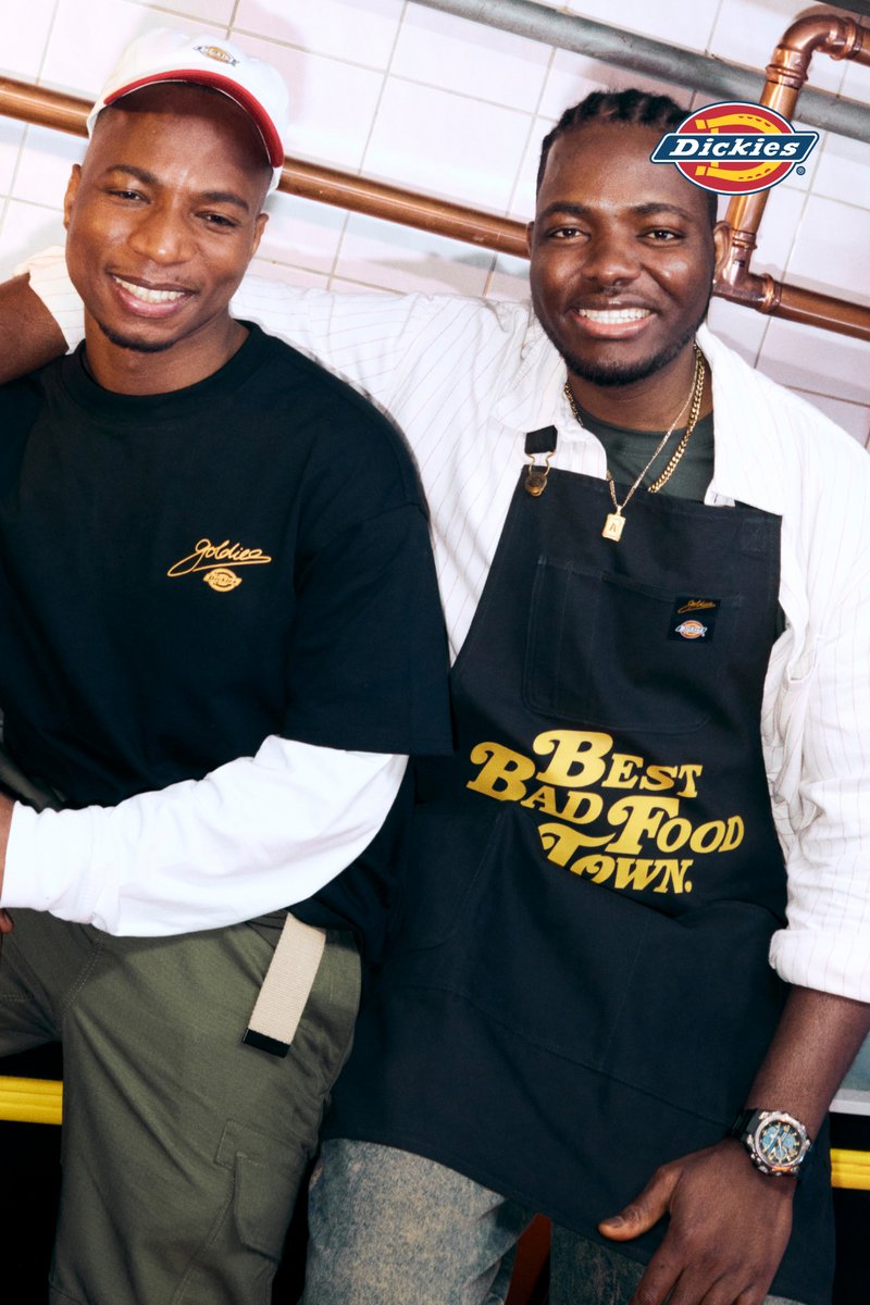 Two smiling men stand side by side, one in a black shirt and cap, the other in a white shirt and apron, in a casual kitchen setting.