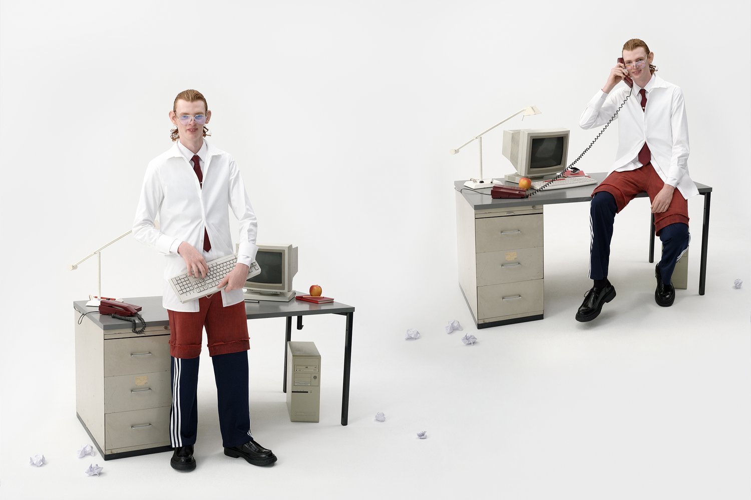 Un joven con una camisa blanca y pantalones cortos rojos sostiene un teclado, de pie al lado de un escritorio con computadoras vintage, una manzana y papeles arrugados.