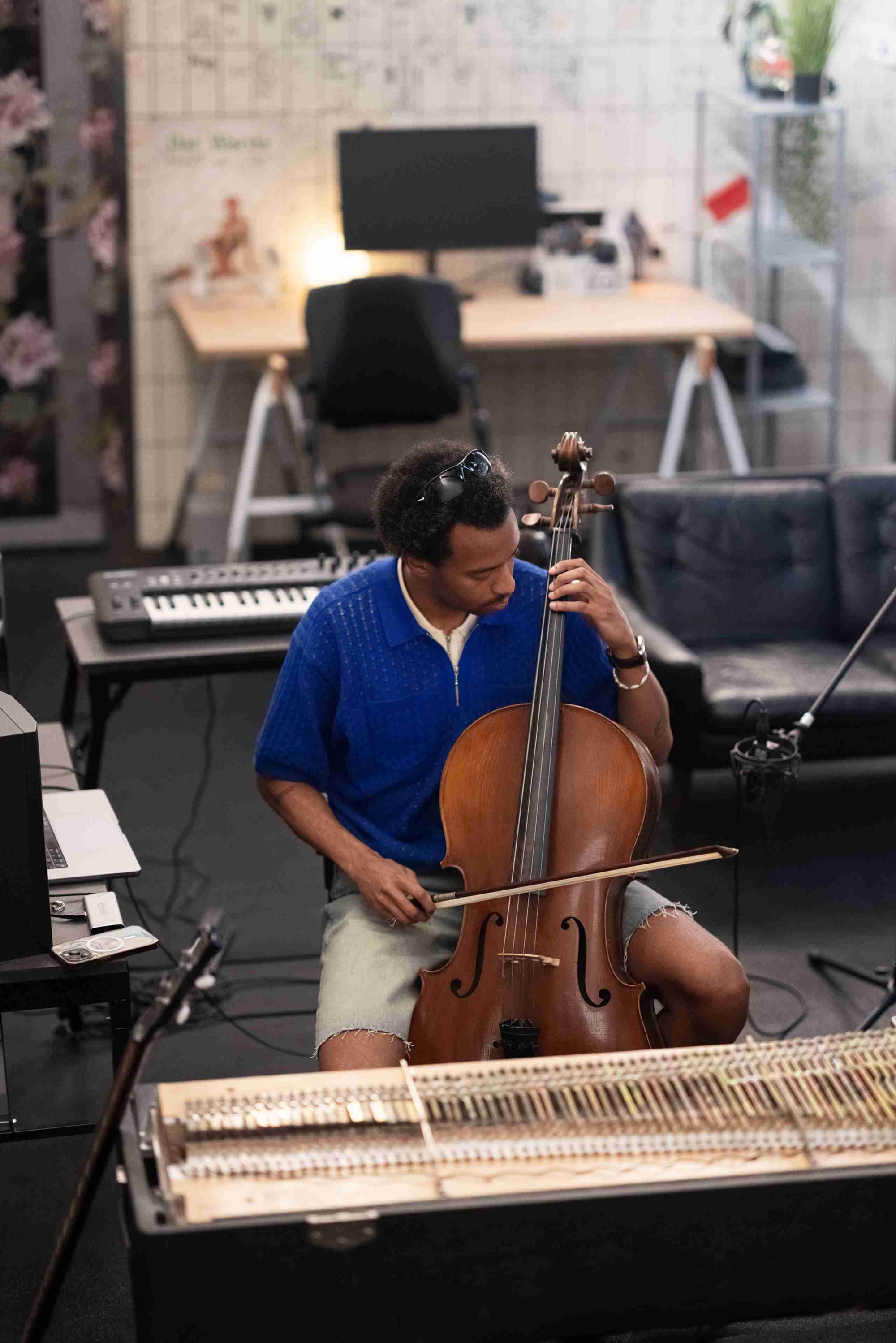 Cellist die een houten cello met een natuurlijke afwerking bespeelt, gekleed in een blauwe kortarmige shirt en denimshorts, in een moderne studio-omgeving.