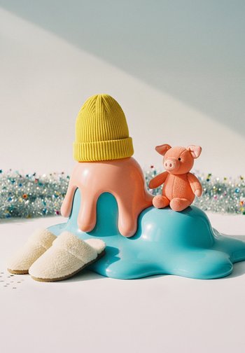 Színes játékok egy teal olvadozó struktúráján, köztük egy sárga bordázott sapka, egy plüss rózsaszín malac, és egy fehér bolyhos papucs világos háttér előtt.