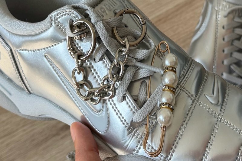 Silberner Nike-Sneaker mit grauen Schnürsenkeln, verziert mit einer silbernen Kette und einer großen goldenen Sicherheitsnadel, die mit Perlen geschmückt ist.
