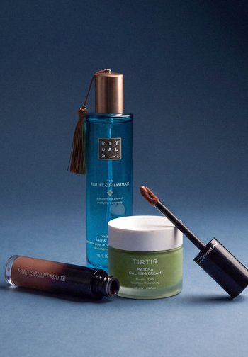 Spray Blue Rituals Hammam bleu, pot vert de crème apaisante Tirtir Matcha, et rouge à lèvres liquide marron MAC avec applicateur sur fond bleu.