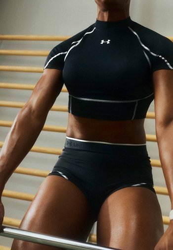 Atleet draagt een zwarte Under Armour crop top en korte broek, houdt een halterstang vast in een sportschool met houten wandrekken op de achtergrond.