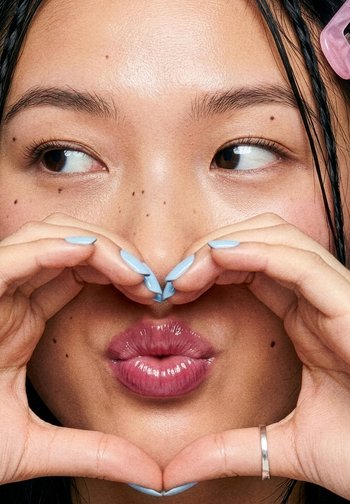 Persona con labbra lucide e capelli intrecciati forma un cuore con le mani, guardando di lato, unghie dipinte di azzurro chiaro.