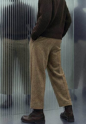 Brune teksturerte bukser med avslappet passform, kombinert med mørkebrune snøreboots. Bakgrunnen har vertikale reflekterende paneler.