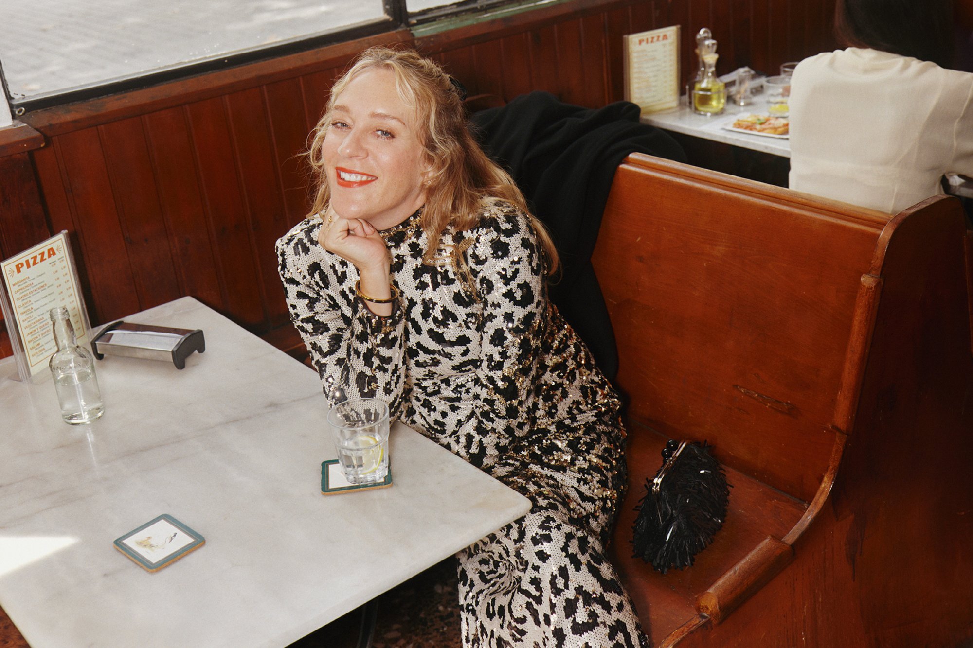 Una donna in un vestito di paillettes sorride mentre poggia il mento sulla mano a un tavolo di ristorante, con un bicchiere d'acqua e un menu visibili.
