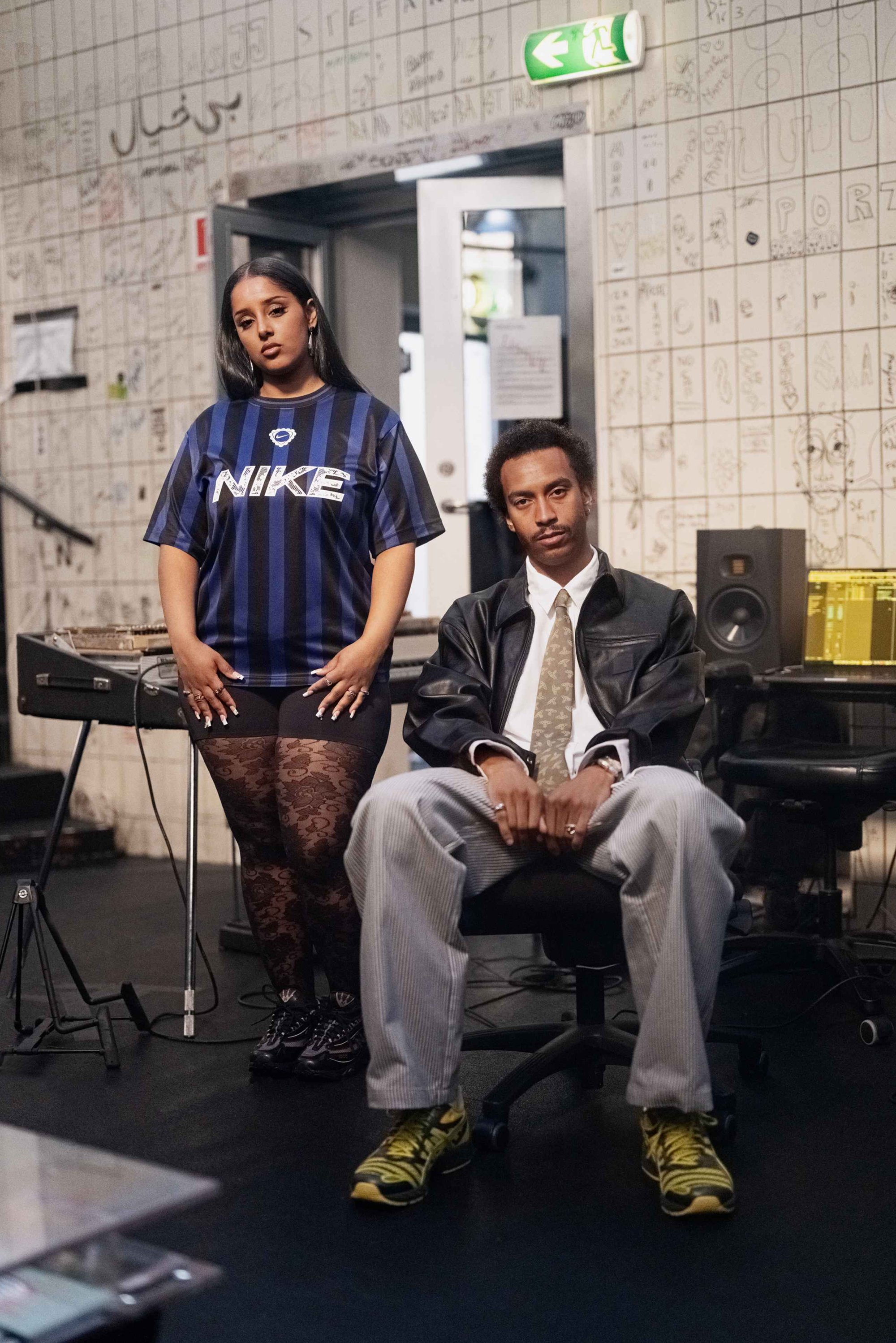 Een vrouw in een blauw gestreept Nike-shirt en zwarte broek met patroon staat naast een man in een zwart leren jack en gestreepte broek die zit.