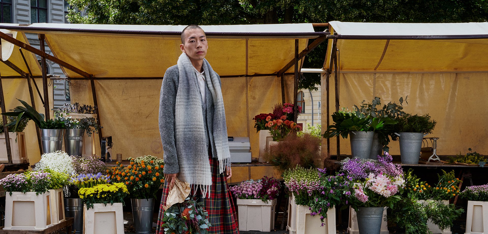 Grey knitted cardigan, beige scarf, and plaid skirt; holding a floral arrangement. Flower market with potted plants and various blooms in the background.