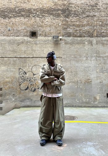 Un homme portant un casque audio, des lunettes de soleil et des vêtements amples couleur kaki se tient les bras croisés dans une cour en béton, à côté de graffitis.
